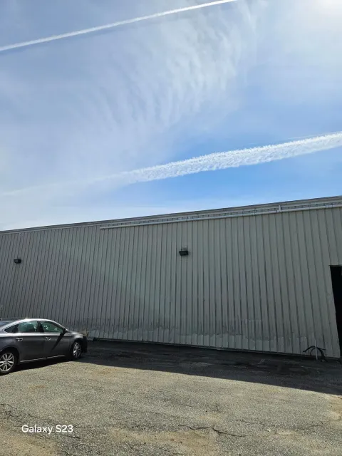 Gray building with a wavy side, car parked on gravel, blue sky with contrails.