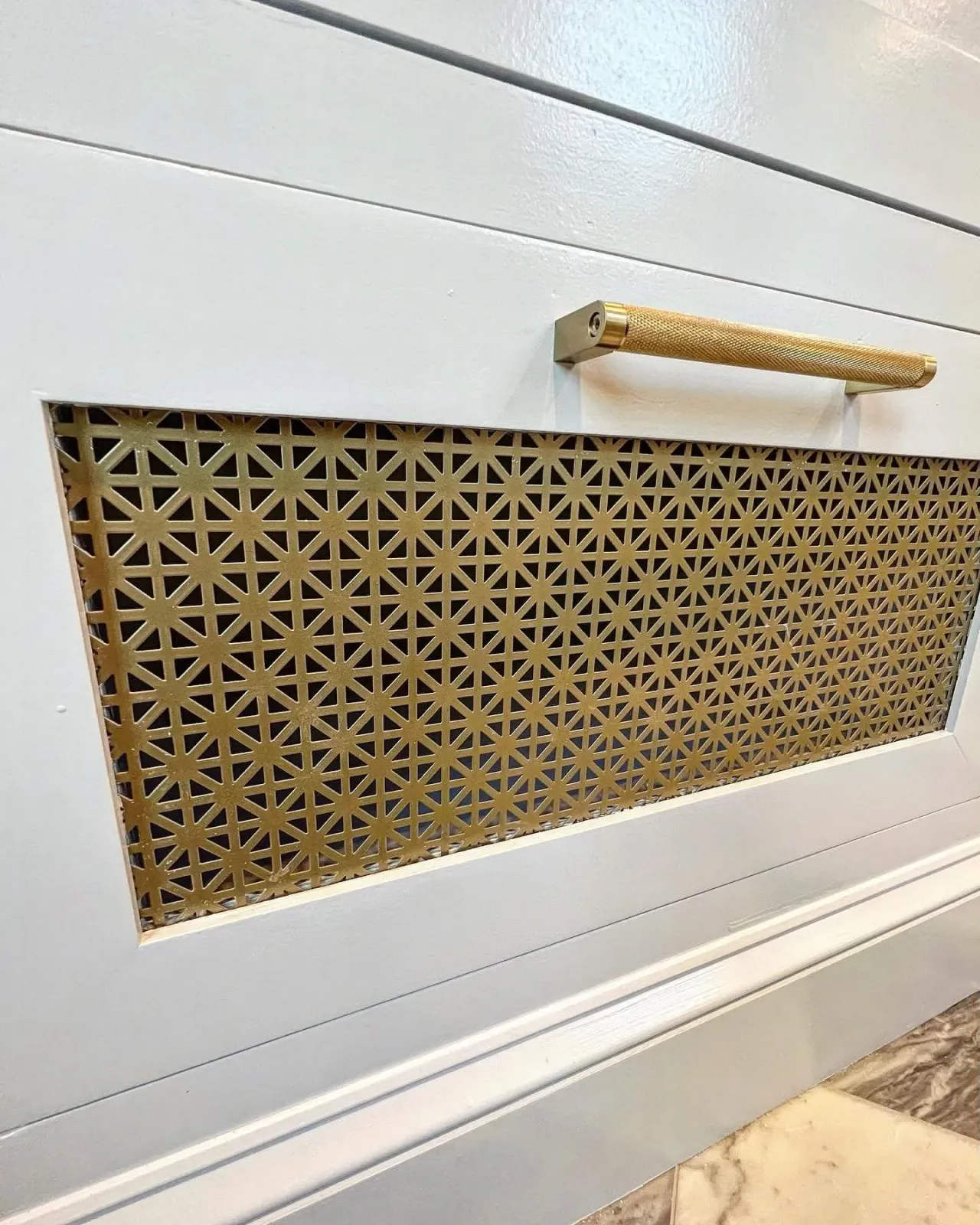 A white panel with a decorative gold grill inserting into it, and a gold handle above the grill.