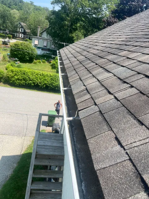 Roof with gray shingles and a gutter, viewed from a ladder beside a driveway and green yard.