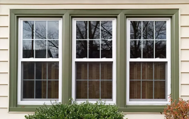 A triple-pane window with white frames and grids, set against light-colored horizontal siding with green exterior trim.