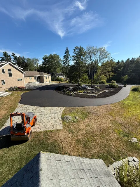 Asphalt driveway curves around a landscaped area with trees, under a blue sky.