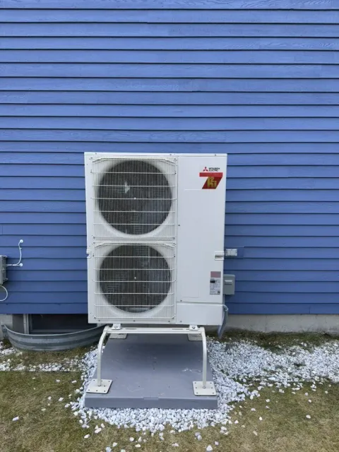 Mitsubishi heat pump unit mounted on a blue house exterior with a gravel base.