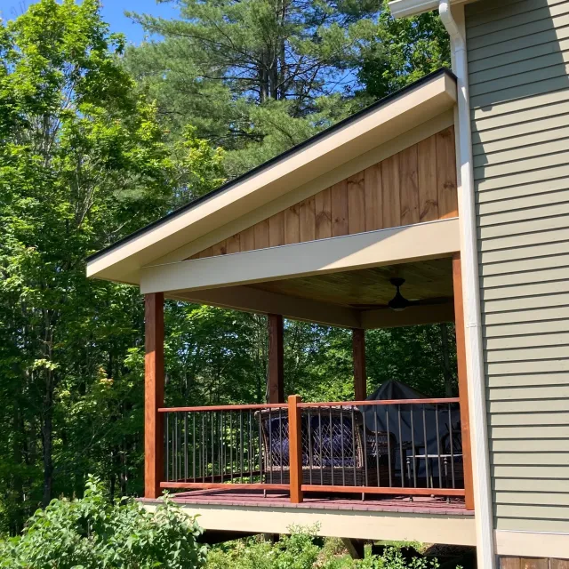 Two-story house with a covered deck with railing, ceiling fan, outdoor furniture, and a grill, surrounded by green leafy trees.