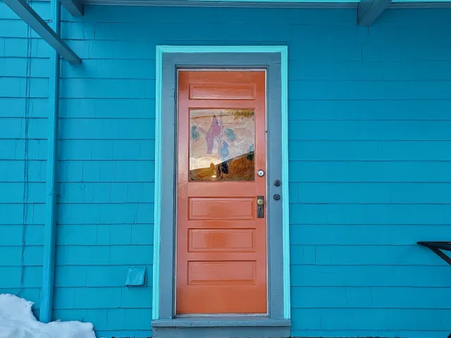 High gloss orange porch door on a colonial in Dorchester, with a decorative window, set in a blue house with horizontal siding, with snow on the ground.