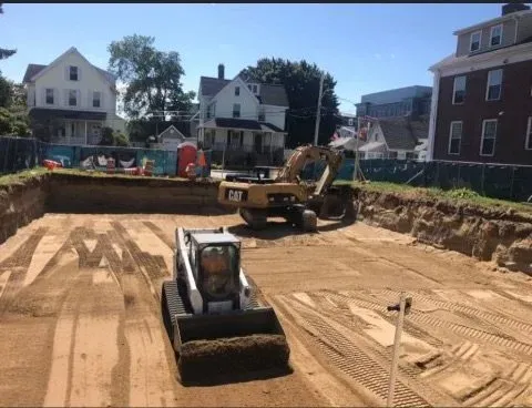 Construction site with an excavator and a small compact roller on a dirt lot, surrounded by residential houses.