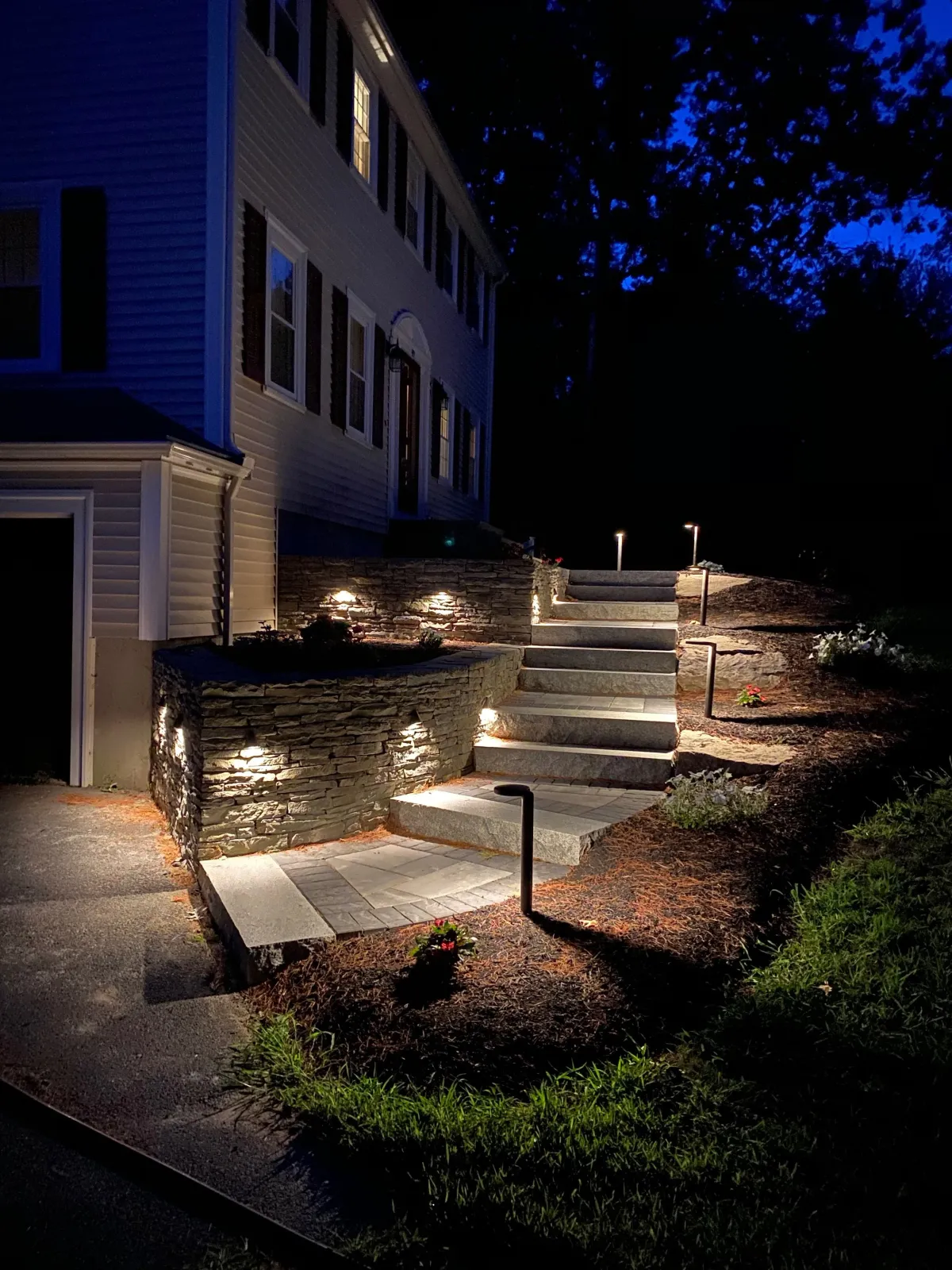 Night view of a tiered outdoor staircase with lighting, leading up to a house with white siding and multiple windows, surrounded by landscaped gardens and flowers.