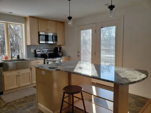 Kitchen with granite countertop island, wooden cabinets, stainless steel microwave, oven, dishwasher, window with outdoor view, glass doors with blinds, and two hanging light fixtures.