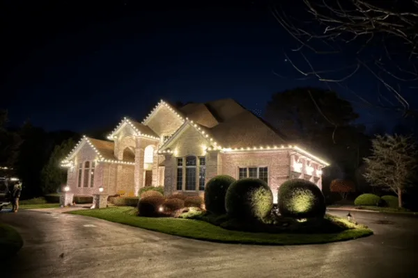 Elegant home illuminated with outdoor lighting and Christmas lighting along the roofline. A dark driveway leads to the well-lit house with manicured bushes in the front yard.