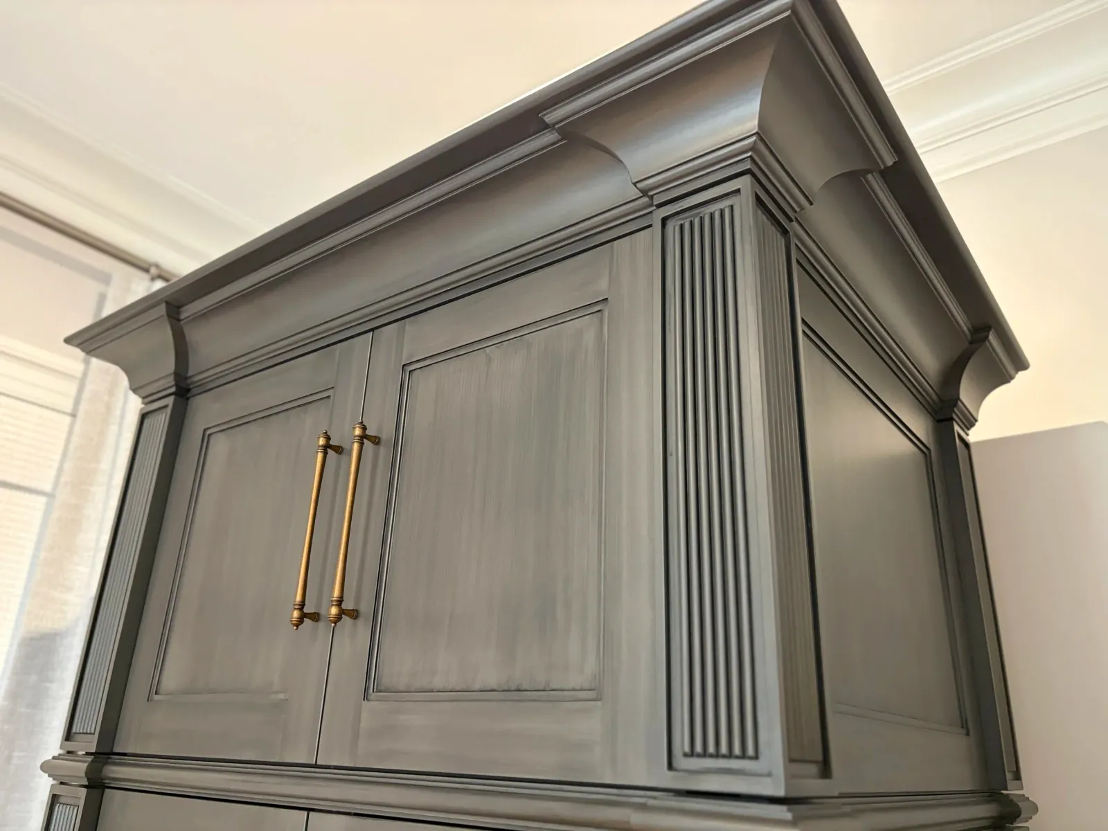 A dark gray wooden cabinet with ornate molding and gold handles, situated in a room with a light-colored ceiling and crown molding.