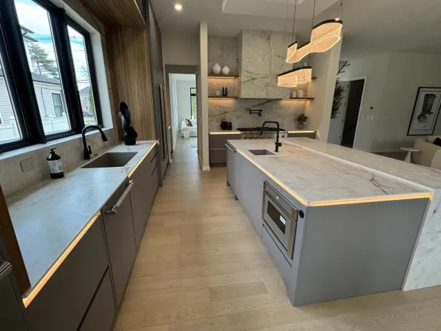 Modern kitchen with gray cabinets, marble countertops, black fixtures, and pendant lighting. Features a large island with a built-in microwave and sink, with view of windows and adjacent living area.