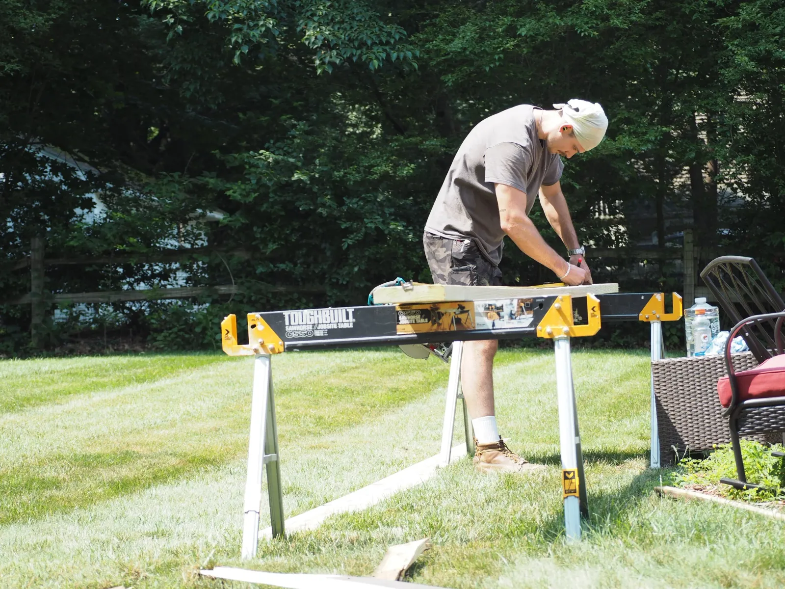 A man wearing a white bandana, gray t-shirt, and camo shorts is cutting a piece of wood on a portable workbench in a backyard with green trees in the background.