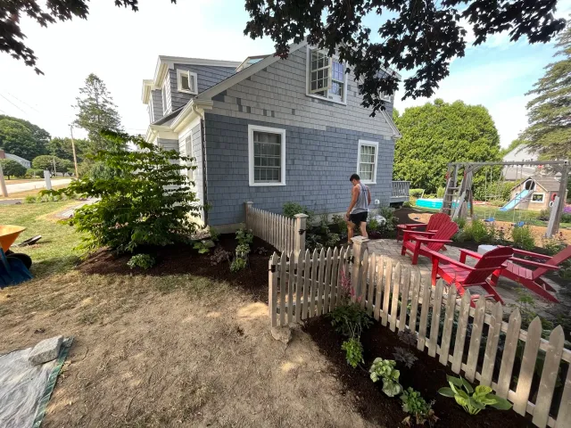 A backyard with a house, garden bed, patio area, and playground equipment. A person is walking on the patio, which has red Adirondack chairs. The yard is landscaped with plants, trees, and a small white fence.