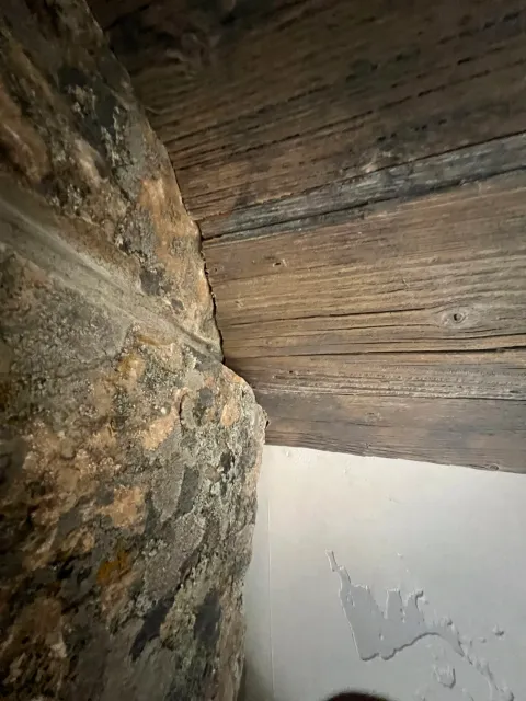 Close-up of a corner where a stone chimney meets a wooden ceiling and a white wall.