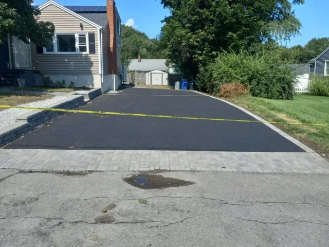 Newly paved asphalt driveway with a gray stone border, beside a house on a sunny day.