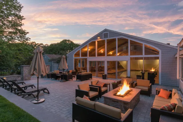 Outdoor patio with fire pit, lounge chairs, and seating area in front of a house with large windows and a porch, at sunset.