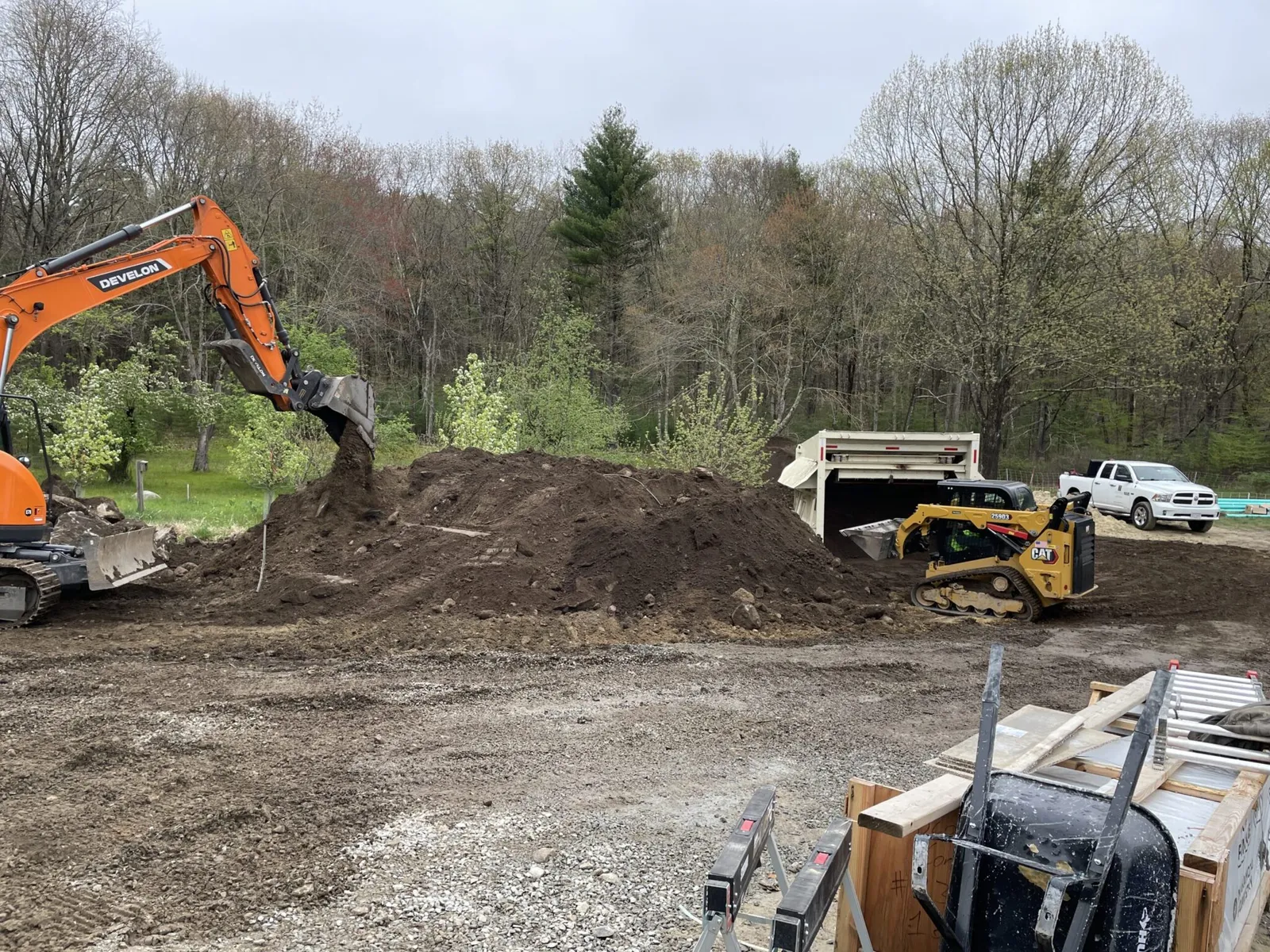 An orange excavator and a CAT skid steer move a large mound of dark topsoil on a graded construction site, with a dump truck, pickup, and early spring woodland visible in the background.