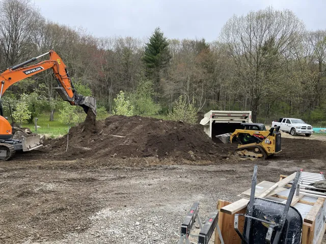 An orange excavator and a CAT skid steer move a large mound of dark topsoil on a graded construction site, with a dump truck, pickup, and early spring woodland visible in the background.