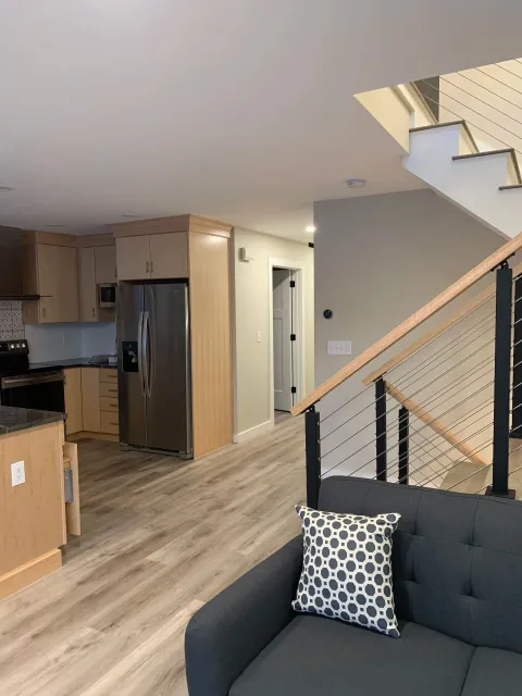 Modern kitchen and living room with stainless steel fridge, light wood cabinets, gray couch, and staircase.