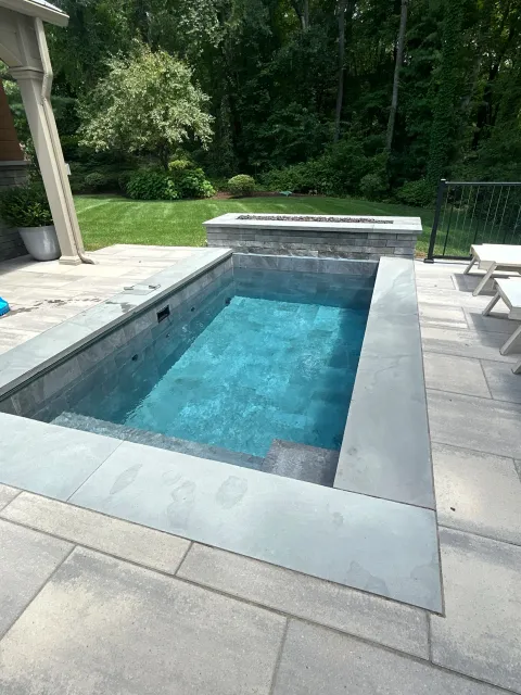 A modern rectangular in-ground swimming pool with light gray stone surround on a patio, adjacent to a grassy yard and surrounded by trees.
