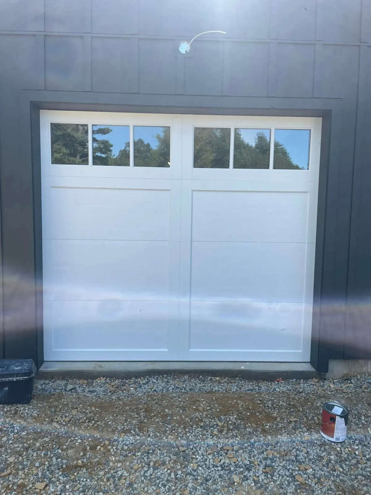 White garage door with windows against a gray building, set in a gravel yard.