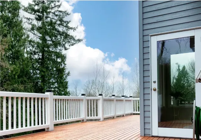 White deck with railing beside gray house and glass door under a blue sky