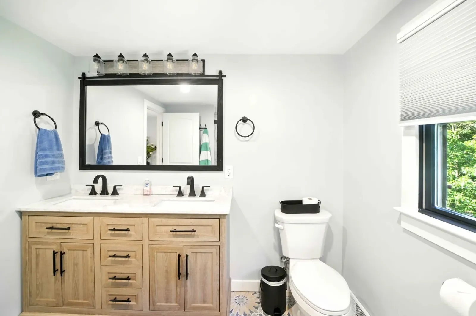 Bright bathroom with wooden vanity, black-framed mirror, toilet, and window with blinds.