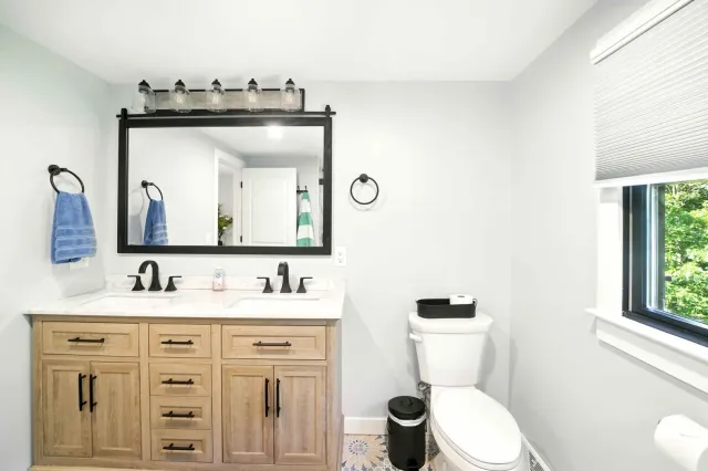 Bright bathroom with wooden vanity, black-framed mirror, toilet, and window with blinds.