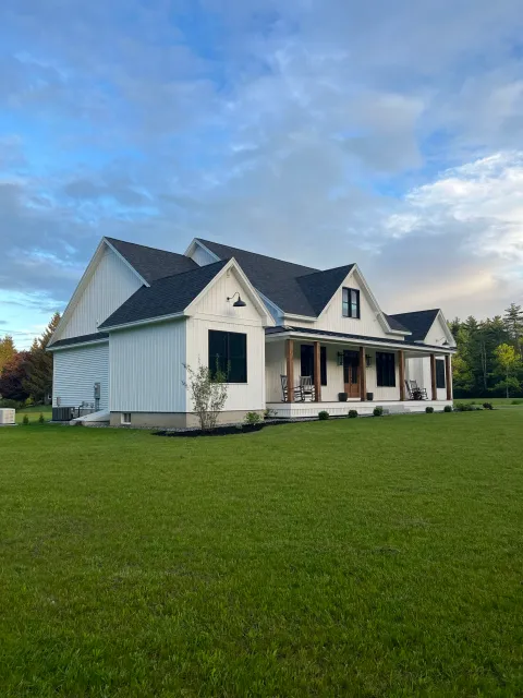 White farmhouse-style house with a large front porch, rocking chairs, black shutters, and surrounded by green lawn and trees under a partly cloudy sky.