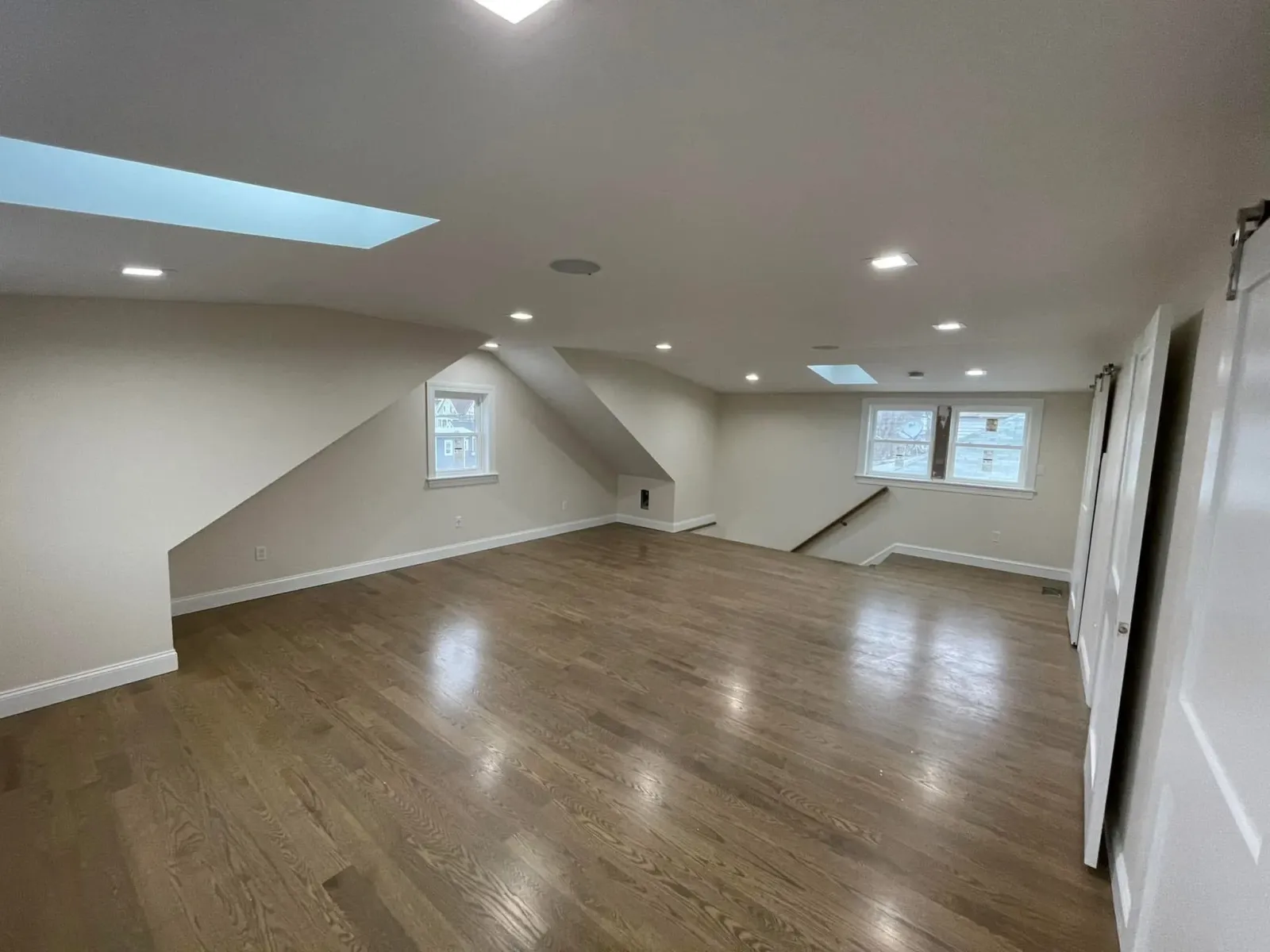 Empty upper-floor attic with hardwood floors, white walls, small windows, recessed lighting, and a staircase with a wooden handrail.
