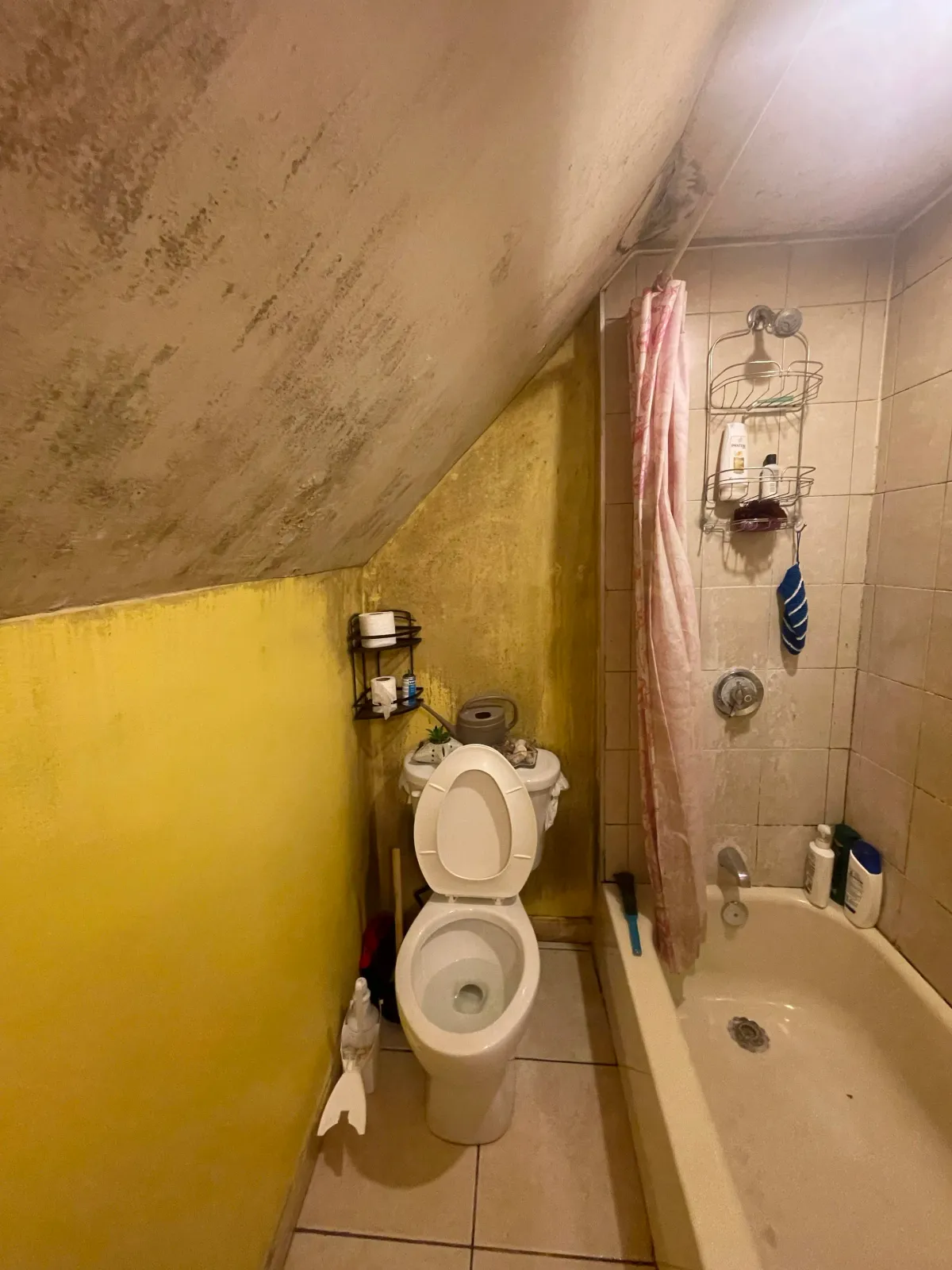A small bathroom with a sloped ceiling, yellow walls, and beige tiled shower area. There is a white toilet with the lid open, a black metal shelf holding toilet paper, and a pink shower curtain. The shower area has a wire rack with toiletries and a b