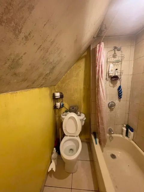 A small bathroom with a sloped ceiling, yellow walls, and beige tiled shower area. There is a white toilet with the lid open, a black metal shelf holding toilet paper, and a pink shower curtain. The shower area has a wire rack with toiletries and a b