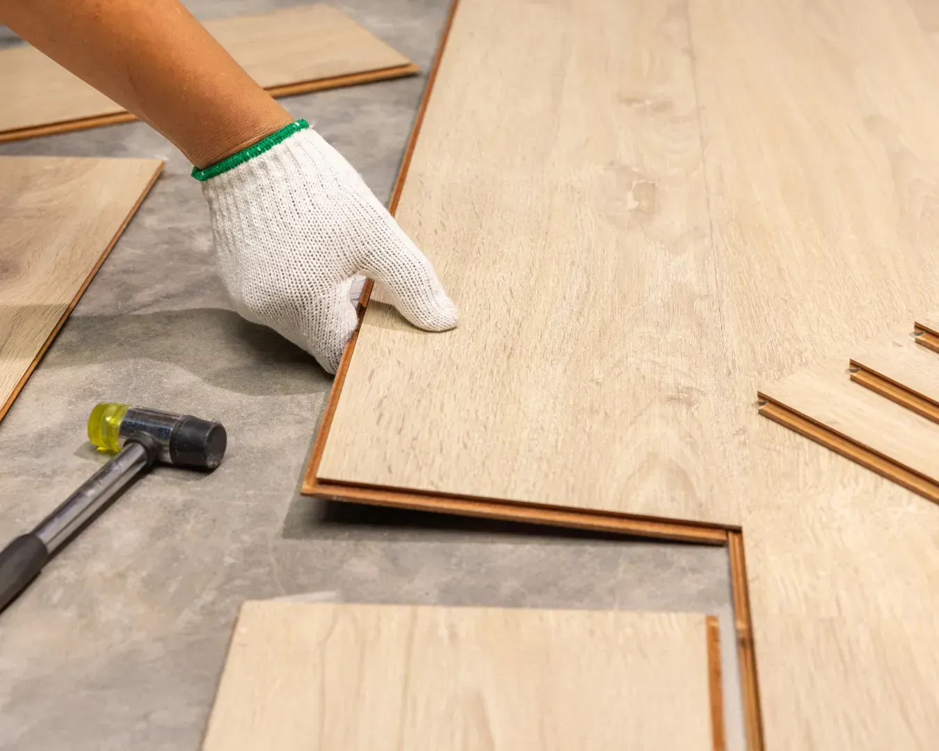 Gloved hand laying wood flooring planks with a hammer nearby