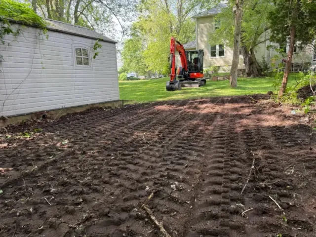 Red excavator grading a dirt yard beside a white shed in a wooded backyard.