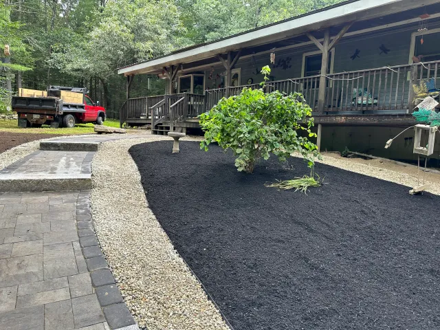 Landscaped yard with black mulch, small tree, stone path, and a rustic porch beside a gravel driveway.