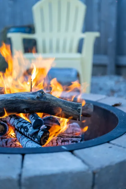 A lit outdoor fire pit with burning logs and flames, a plastic yellow Adirondack chair in the background, and a wooden fence.