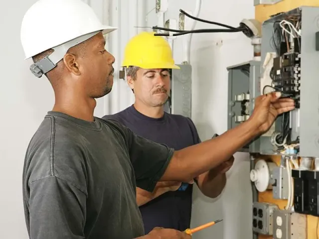Two men wearing hard hats are working on an electrical box