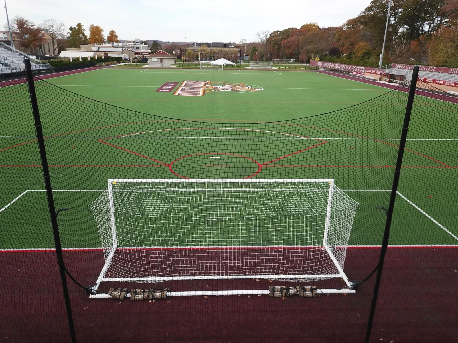 Empty soccer field with goal in foreground, enclosed with black netting, and a college logo in the center of the field, surrounded by bleachers, trees with fall foliage, and a score banner for Rhode Island College.