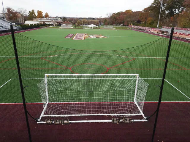 Empty soccer field with goal in foreground, enclosed with black netting, and a college logo in the center of the field, surrounded by bleachers, trees with fall foliage, and a score banner for Rhode Island College.