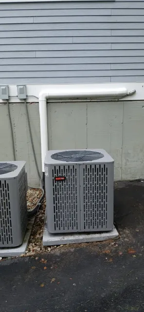 Two gray outdoor HVAC condenser units sit on concrete pads against the side of a building with light gray siding.