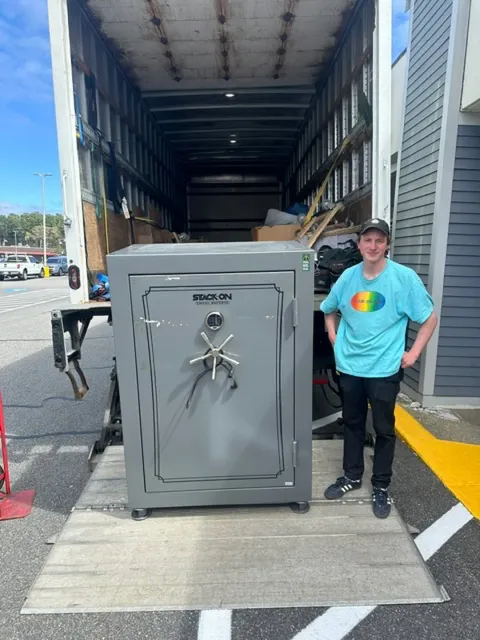 A person standing next to a large gray safe with a wheel lock, in the back of a moving truck, outside a building.