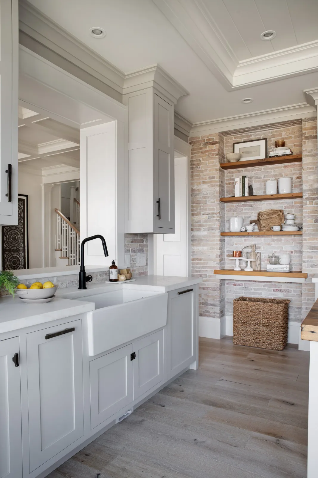 Hardwood floor installation in a Duxbury kitchen with white cabinets, a black faucet, a brick wall with open wooden shelves, and light wood flooring.
