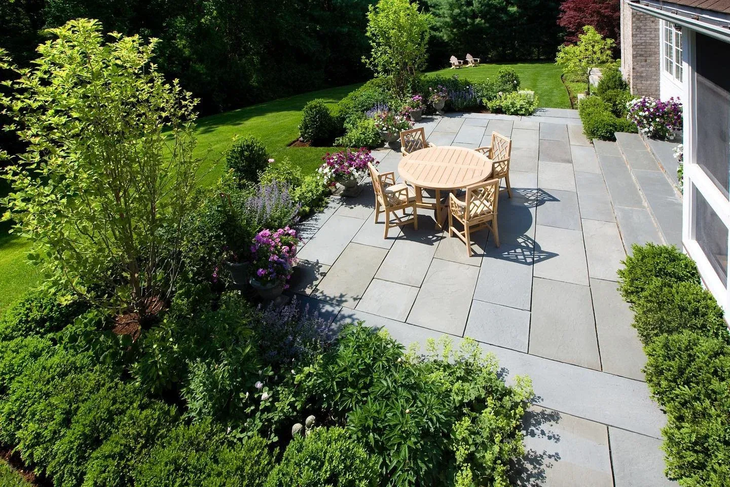 A backyard patio with a round wooden table and six chairs, surrounded by lush green bushes and colorful flowering plants, with a well-manicured lawn and trees in the background.