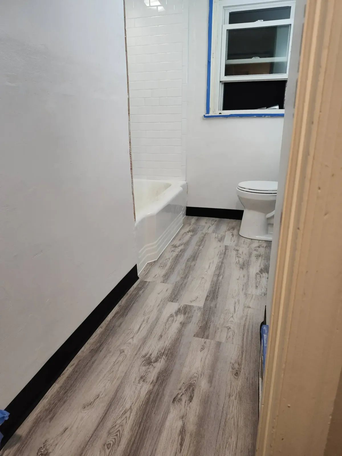 A bathroom with light gray wood-look flooring, white walls, a bathtub with white tile, and a white toilet.