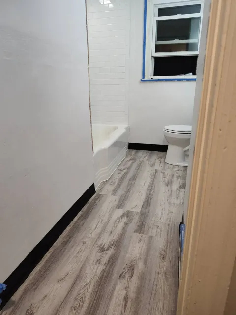 A bathroom with light gray wood-look flooring, white walls, a bathtub with white tile, and a white toilet.