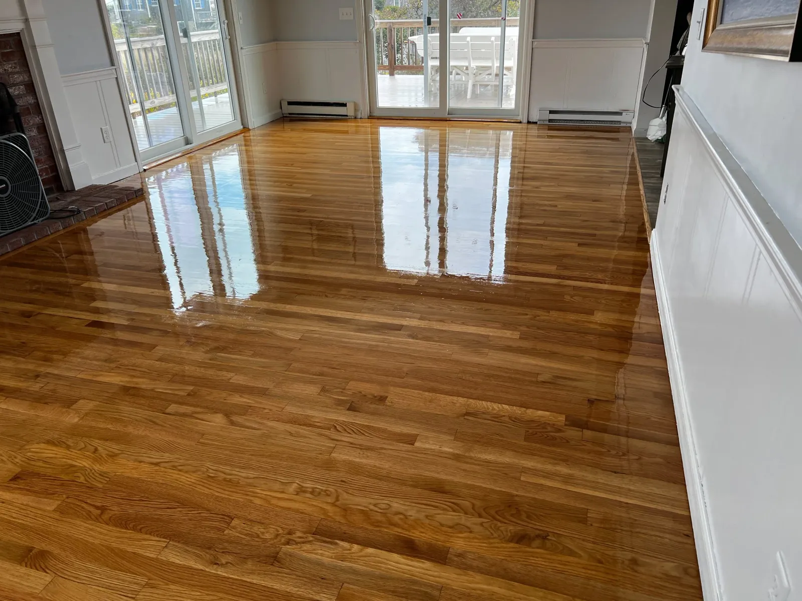 A room with freshly polished hardwood floors reflecting sunlight, with sliding glass doors leading to a deck with outdoor furniture.