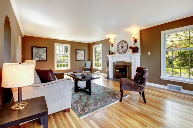 Living room with brown and beige walls, three large windows, white fireplace mantel, hardwood floors, and furniture including a beige sofa with dark red pillows, a brown armchair, and a dark wood coffee table with decorative items. Wall art and a large clock are above the fireplace.