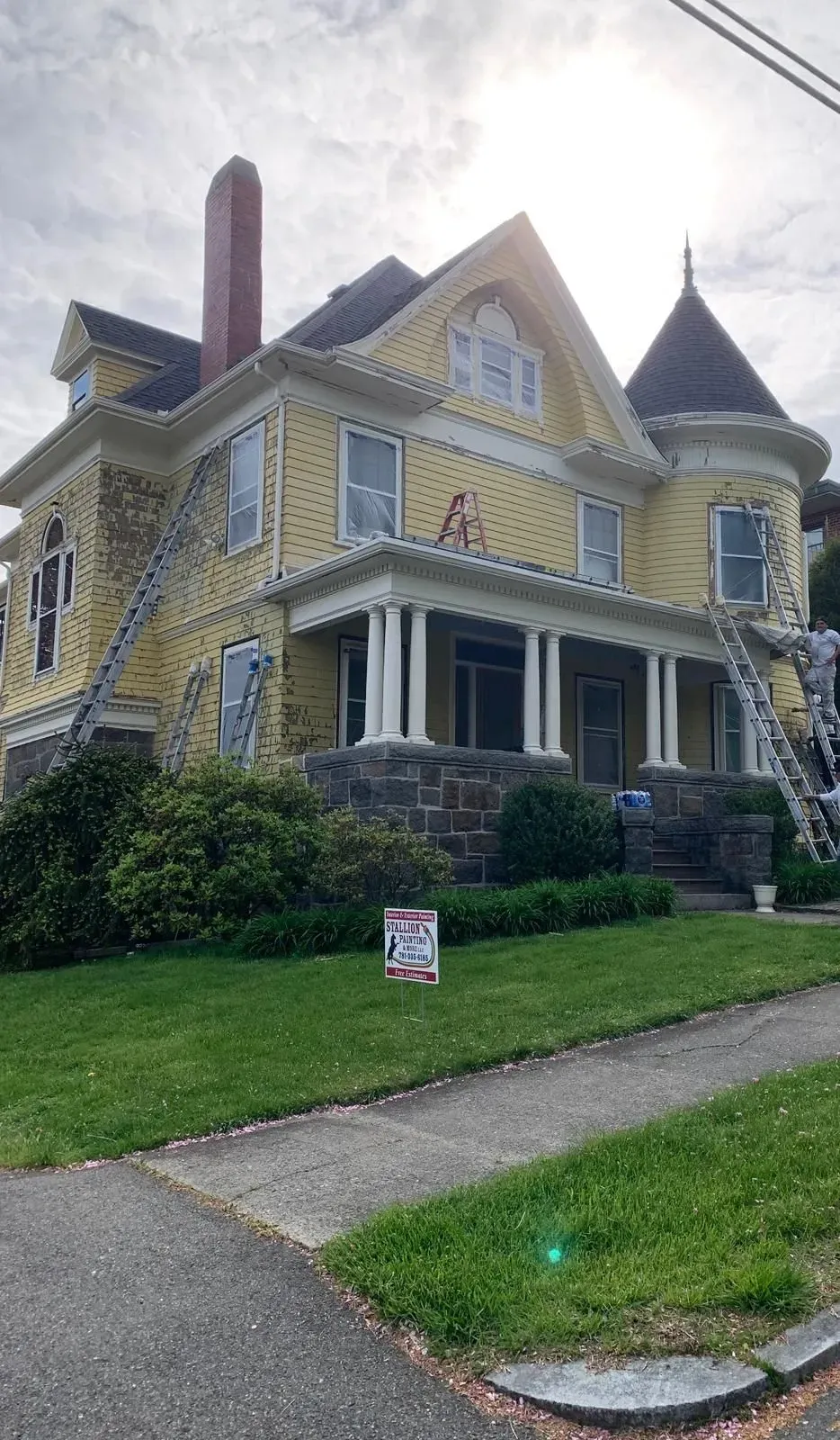 Victorian yellow house with porch, turret, and stone steps on a grassy corner lot