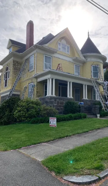 Victorian yellow house with porch, turret, and stone steps on a grassy corner lot