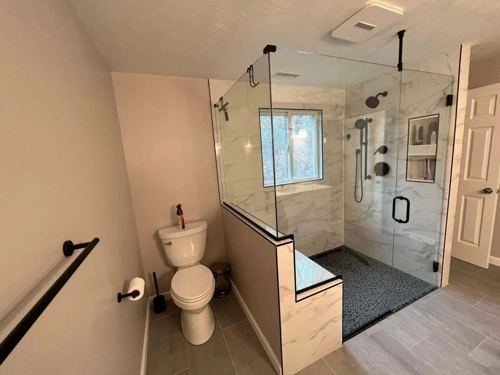 Modern bathroom with toilet, glass walk-in shower, and a black towel bar near a window.
