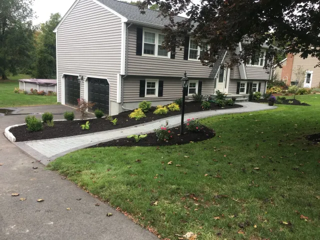 Front yard with a paved walkway, landscaped with small shrubs, flowers, and mulch, leading to a gray house with black shutters and garage doors, surrounded by green grass and trees.